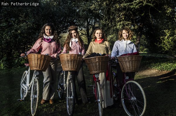  Four women stand in alongside their bicycles, all of which have wicker baskets
